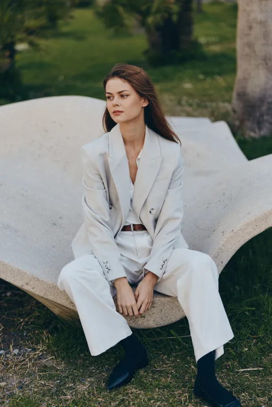 Confident business woman in tailored light gray suit sitting outdoors, exuding focus and elegance amidst nature, showcasing professionalism and style.