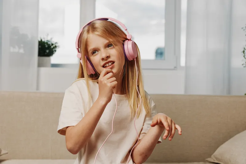 Blonde girl with pink headphones listens to music thoughtfully while sitting on beige couch near window in bright room. Concept of childhood, relaxation, and modern technology use.
