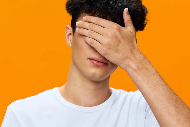 Young man covering face with hand against bright orange background, wearing white t-shirt, casual style, emotional expression, isolated studio portrait.