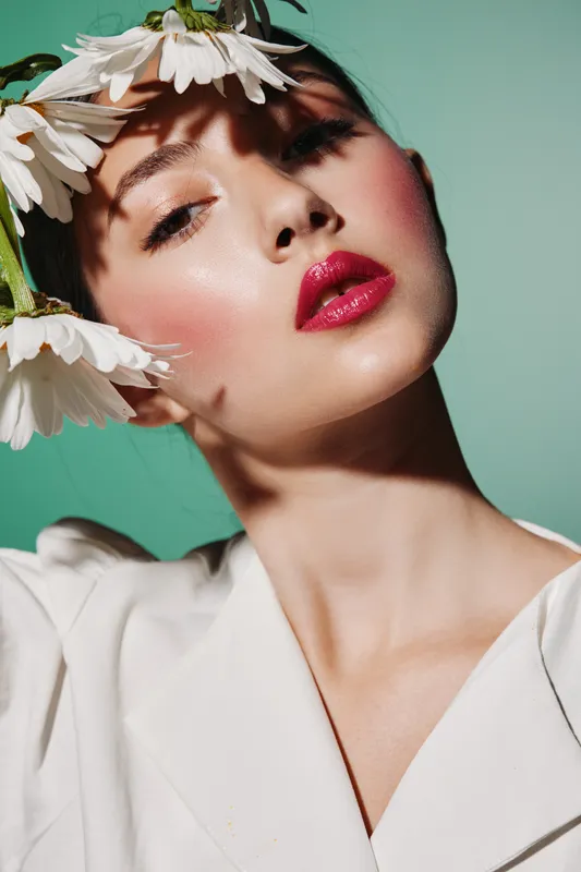 Portrait of a female model wearing vibrant makeup and a beige top. Daisies cast artistic shadows on her face against a green background. Fashion beauty concept in natural light.
