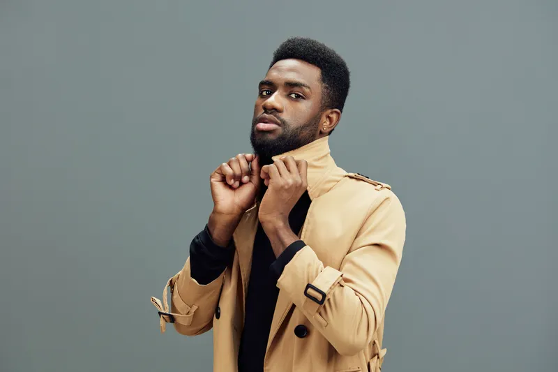 Stylish young Black man in a beige trench coat, confidently adjusting his collar against a gray background, exuding modern elegance and self assurance