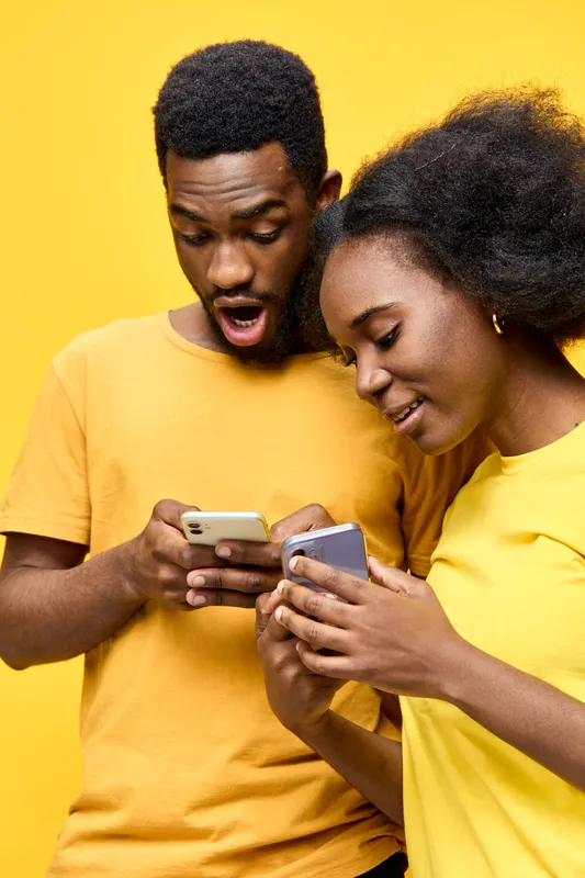 Happy young couple excitedly using smartphones against a bright yellow background, sharing joyful moments together