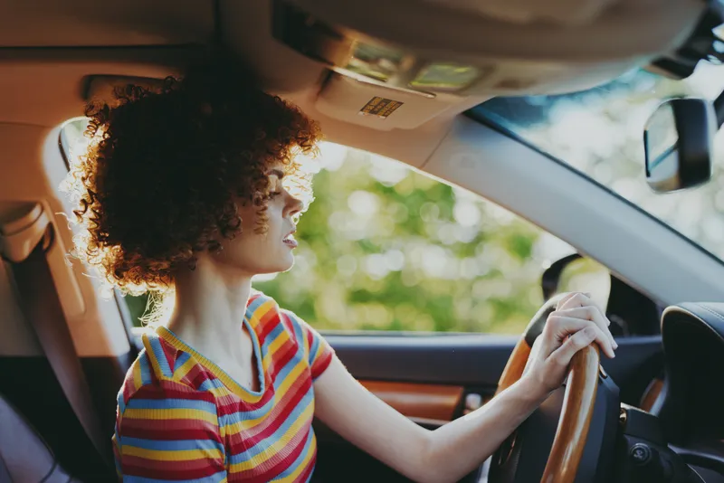 Young woman with curly hair drives a car in daylight, wearing a colorful striped shirt, expression focused and calm as she steers along a sunlit street.