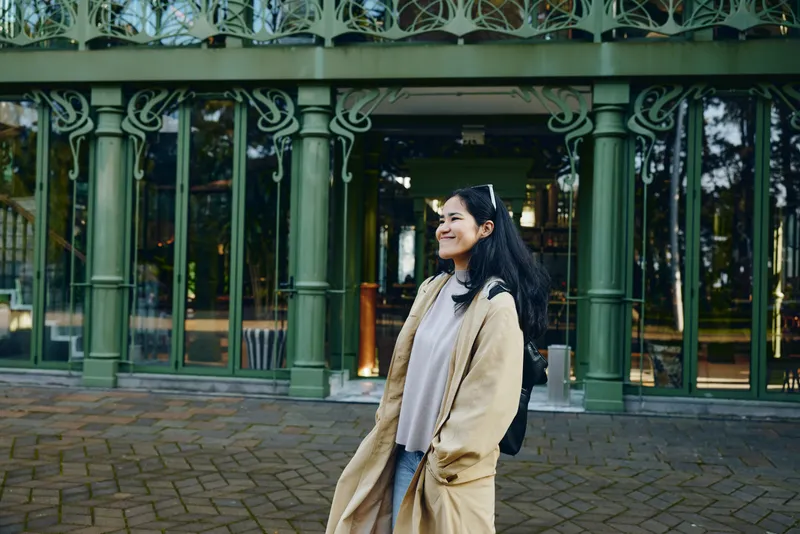Woman in a beige coat with backpack smiling outside a green ornate building, casual street style, urban setting, fashion photography for lifestyle and travel