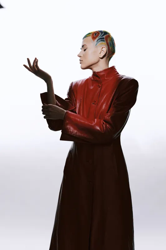 Stylish woman with colorful braided hair wearing a long red leather coat poses gracefully against a clean white background, showcasing modern fashion and unique style.