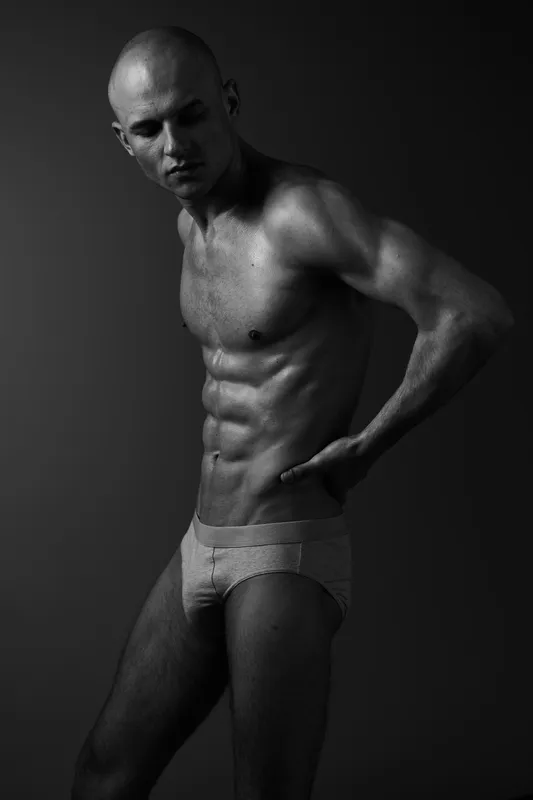 Black and white portrait of a muscular man posing confidently, showcasing his body. The image captures strength and determination in a minimalist style.