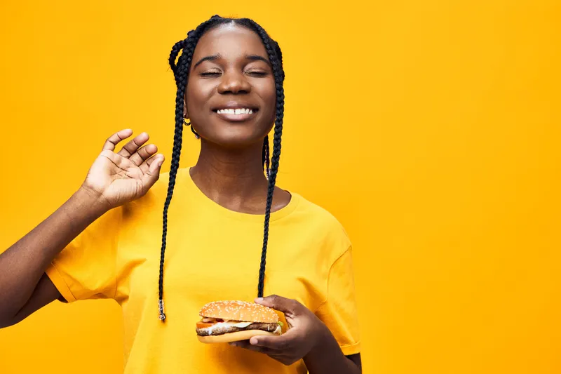 Smiling young African girl enjoying a delicious sandwich against a bright yellow background, expressing joy and satisfaction in a casual summer style