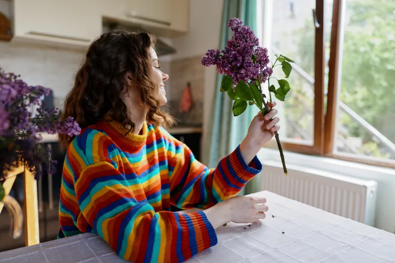 Young woman in a colorful striped sweater sits at a dining table indoors, admiring a lilac bouquet as warm sunlight streams through a sunlit window nearby, creating a cozy home moment.