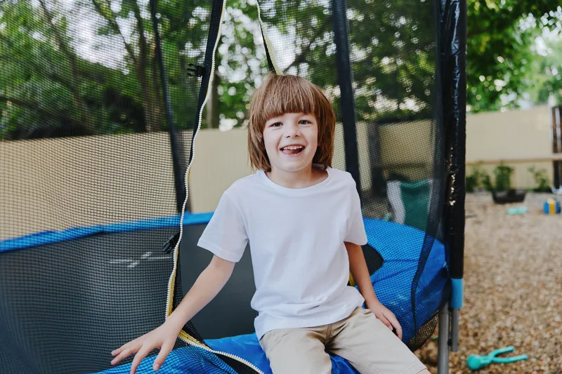 Cheerful boy enjoying a sunny day on a trampoline, surrounded by greenery and a playful atmosphere. Happiness radiates from his smile and carefree spirit.