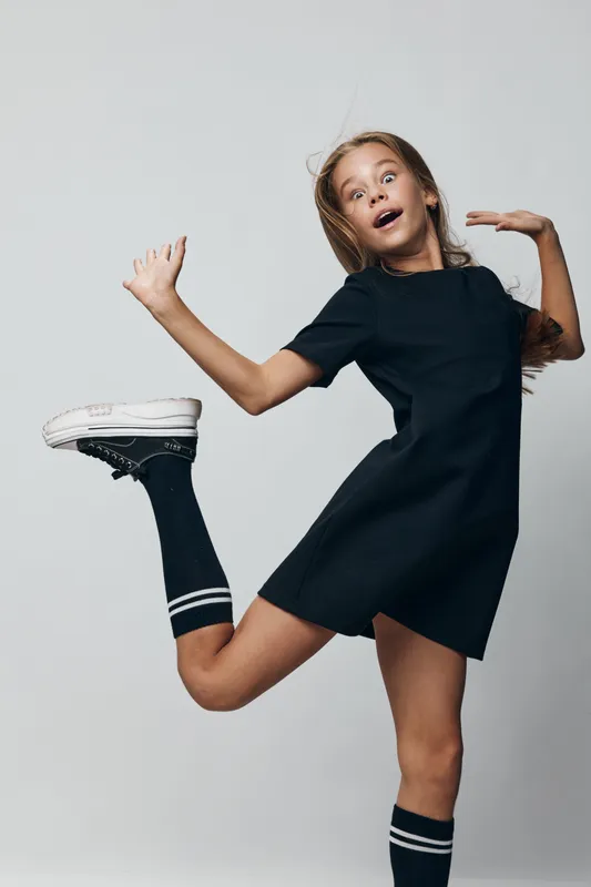 Girl jumping with energy in dark dress and high socks on grey background, expression of excitement and movement captured in studio light.