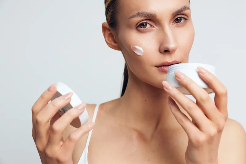 Young woman applying white face cream on cheek holding open jar skincare concept soft natural makeup healthy skin beauty treatment close-up portrait on light background.