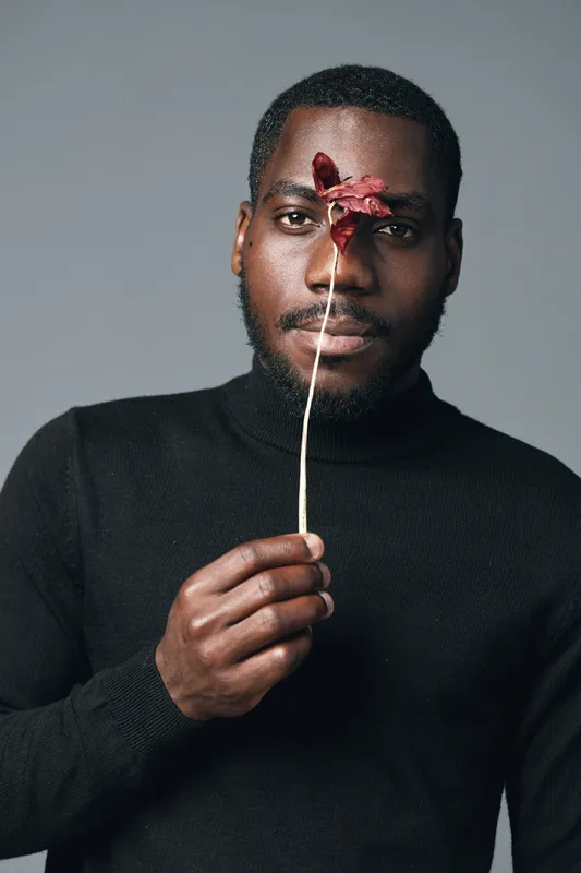 African man with dark skin holding dried red flower in front of face wearing black sweater against gray background creative conceptual visual style portrait with thoughtful expression