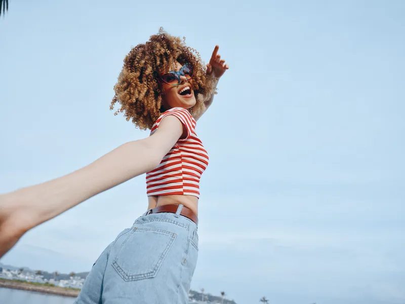 Joyful woman laughing and spinning outdoors, wearing a striped crop top and jeans, reaching toward the camera against a bright blue sky on a sunny day.