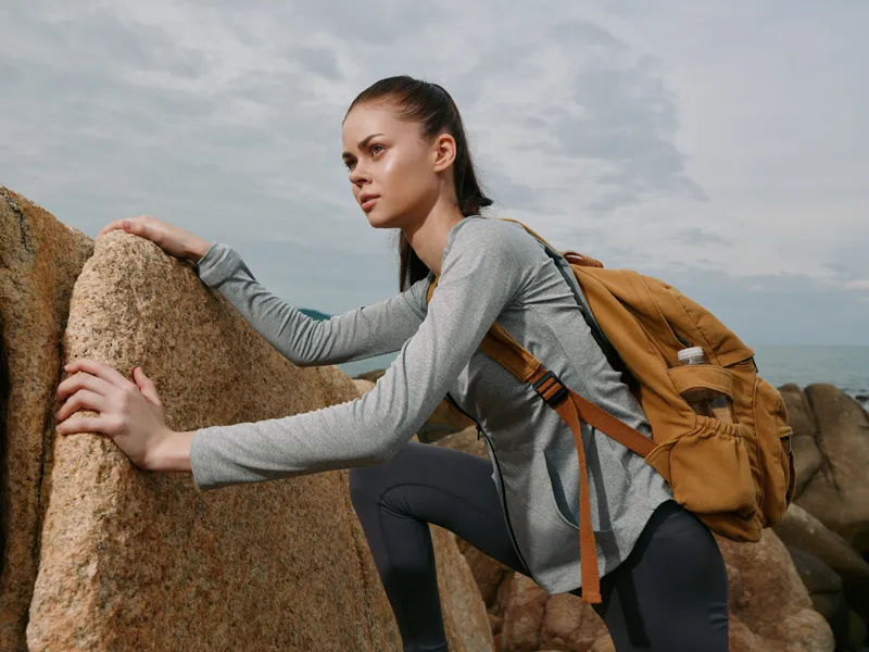 Young female hiker in activewear climbs rocks by the sea, showcasing determination and adventure in a natural landscape