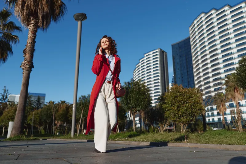 Young woman in red coat walking outdoors in urban park with modern buildings and palm trees. Sunny day with clear blue sky, casual fashion and confident mood.