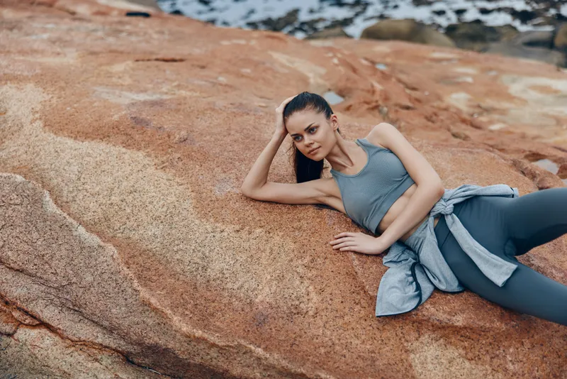 Fitness model posing on rocky beach in activewear, showcasing sporty lifestyle and natural beauty against coastal backdrop