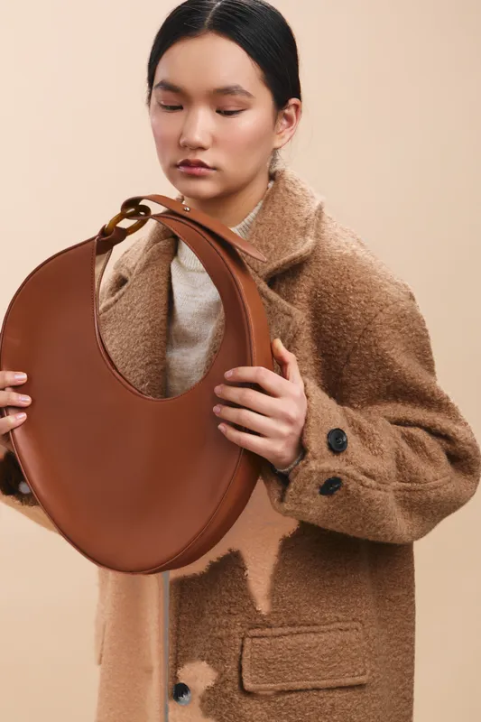 Asian woman holds a round brown bag in a soft studio setting, wearing a cozy tan coat. The scene conveys calm fashion, warmth, and contemporary accessory styling.