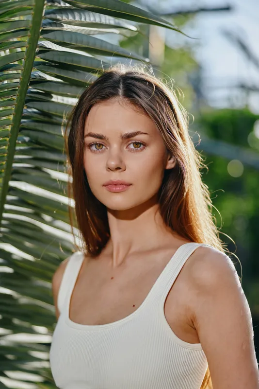 Young woman with long brown hair wearing white tank top standing near palm leaves in soft natural sunlight outdoors, showcasing serene expression and casual style.