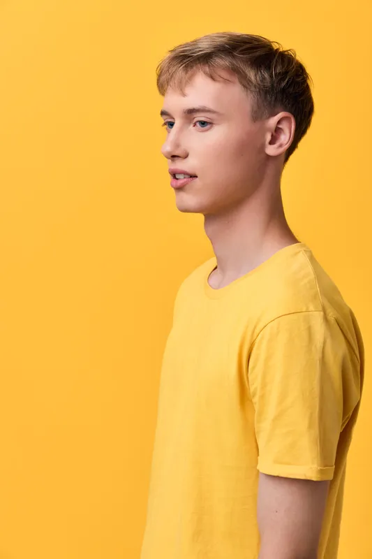 Boy in a yellow shirt stands in profile against a bright yellow backdrop, clean studio shot with a minimal scene, emphasizing color, shape, and calm mood for fashion or lifestyle use.