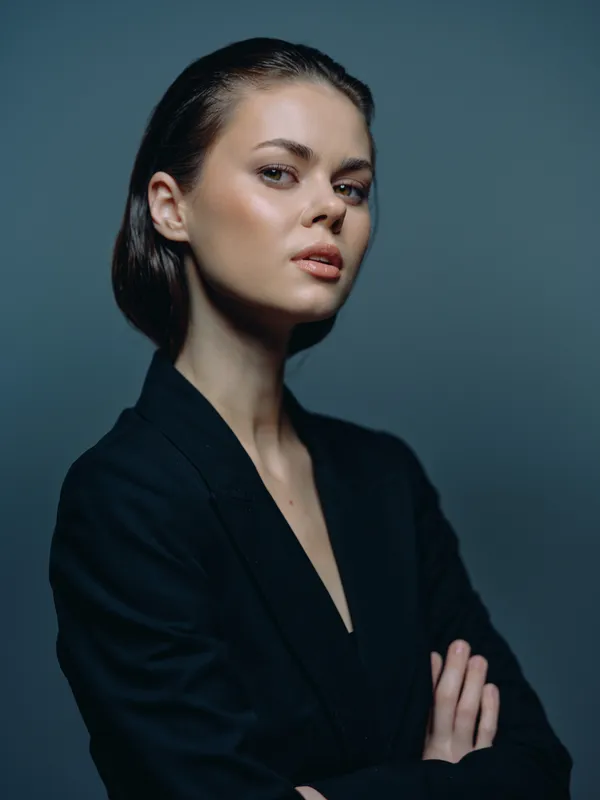 Confident woman in black blazer with folded arms against dark background, portrait of poised young adult, stylish and serious expression.