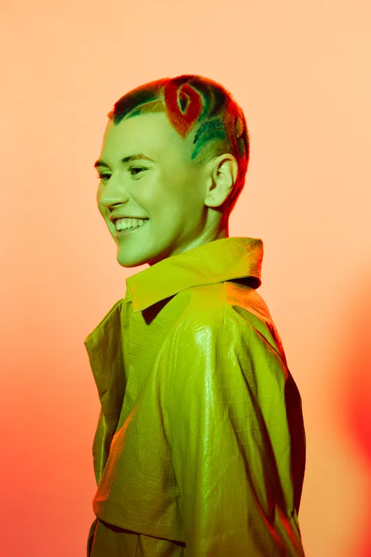 Young woman with colorful hairstyle and bright yellow coat smiling against orange background, modern fashion, creative style, vibrant mood portrait.