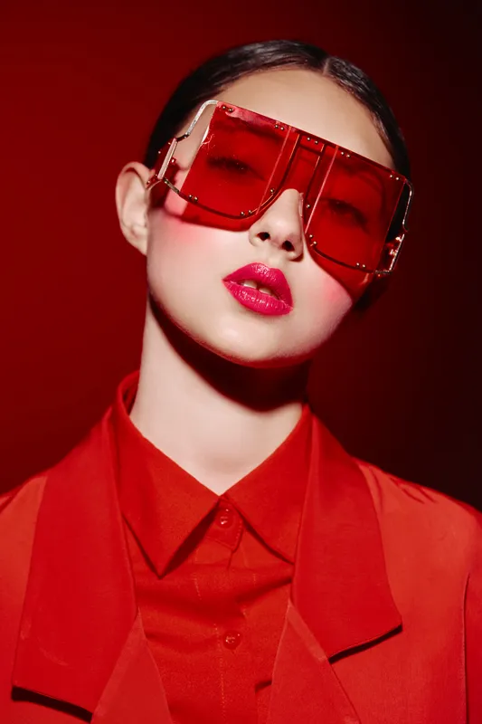 Young woman with bold red lipstick wearing large red sunglasses and vibrant red clothing. Editorial fashion beauty portrait with intense monochrome red studio background.