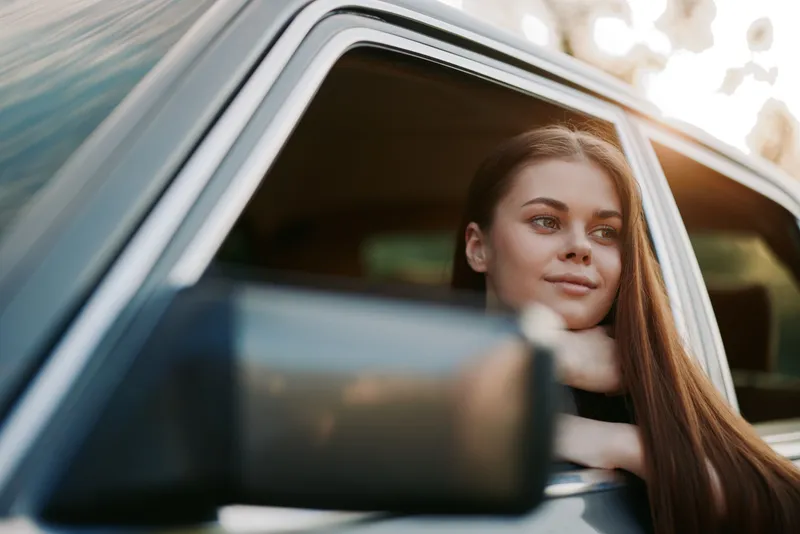 Young woman enjoying a sunny day, looking out of a vintage car window, exuding a thoughtful and relaxed expression, with natural lighting and a warm color palette.