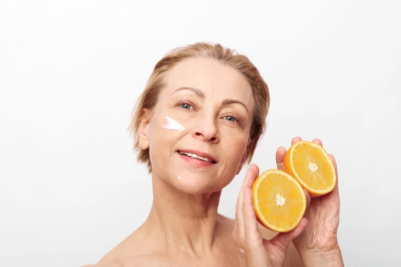 Mature smiling woman with natural skin holds fresh orange halves near face with white cream on cheek. Concept of skincare, healthy lifestyle, natural beauty, and citrus fruit wellness.