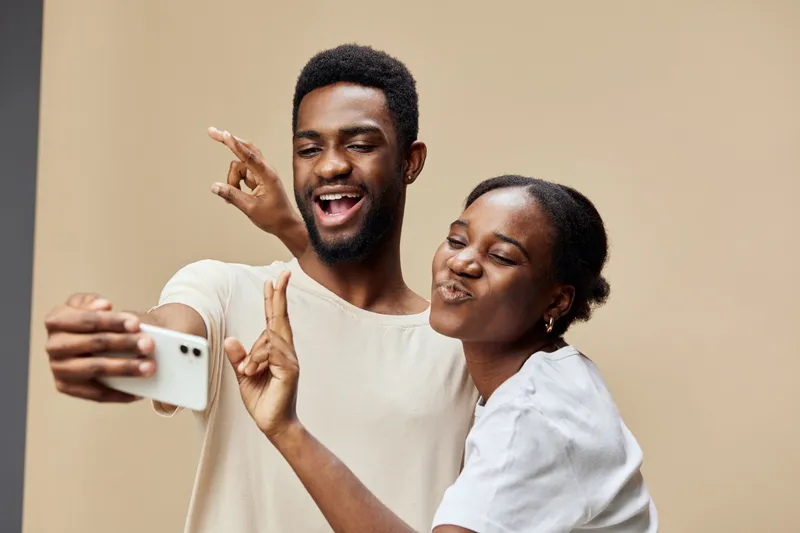 Happy young couple taking selfies together, capturing joyful moments with playful expressions against a minimalist beige background