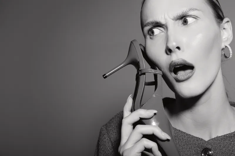 Confused white woman with pale skin holding a high heel shoe near her ear as if it is a phone, studio portrait with dramatic expression and grey background.