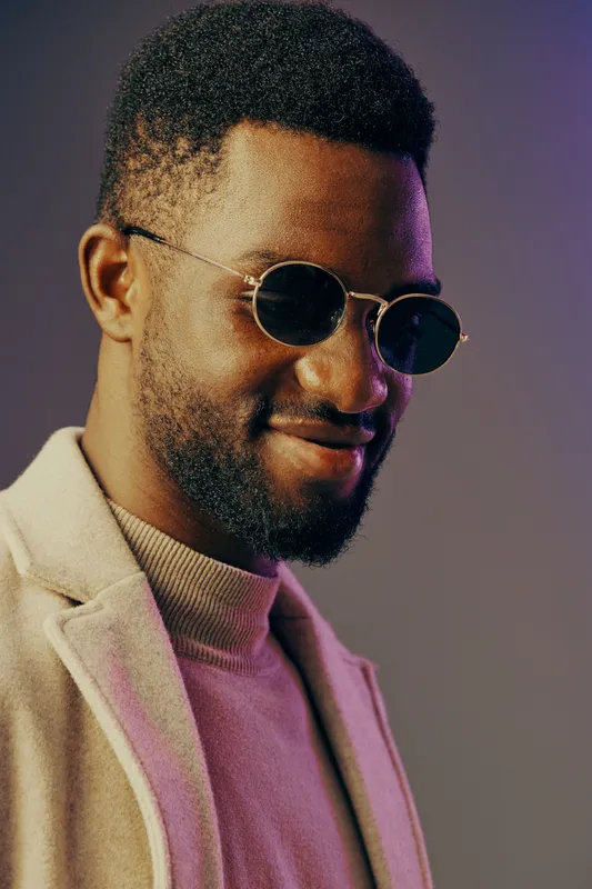 Stylish African man wearing sunglasses and a fashionable coat smiles confidently against a pastel colored background, capturing modern elegance and confidence