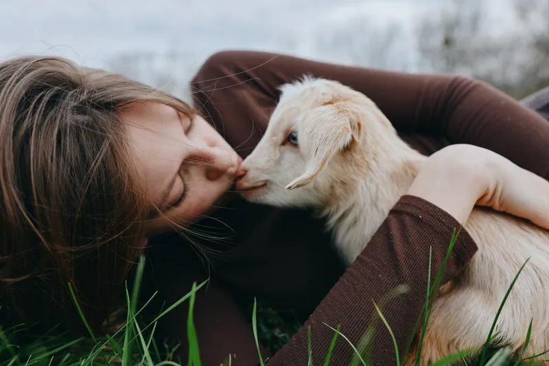 Lifestyle moment of a girl lying on grass, kissing a gentle goat kid, showing tenderness and a bond with animals in an outdoor setting, calm mood, soft light, candid portrait of companionship
