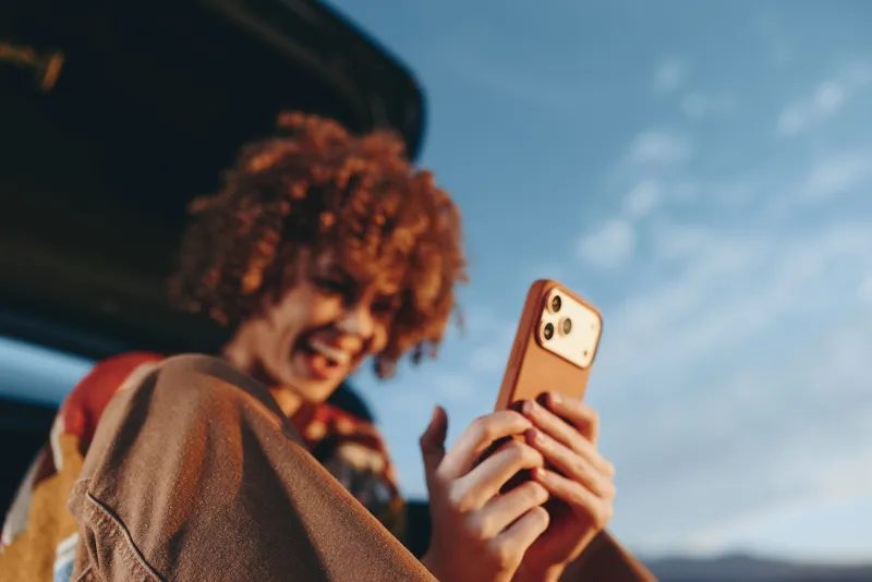 Woman with curly red hair holds a smartphone for a selfie outdoors, casual lifestyle portrait, technology moment, bright sky background, she smiles naturally at the phone today
