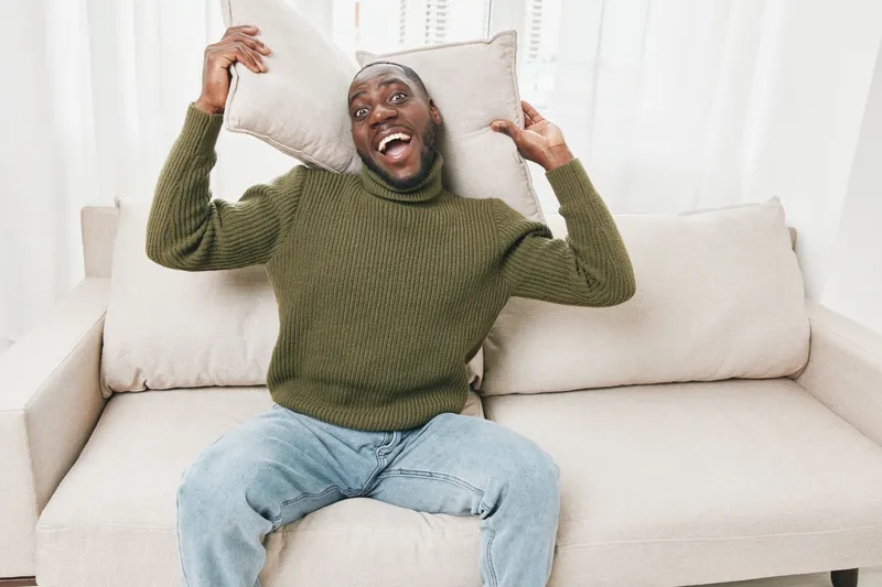 Happy African American man in green sweater smiling with pillows on white couch indoors, showcasing joy and comfort in modern lifestyle