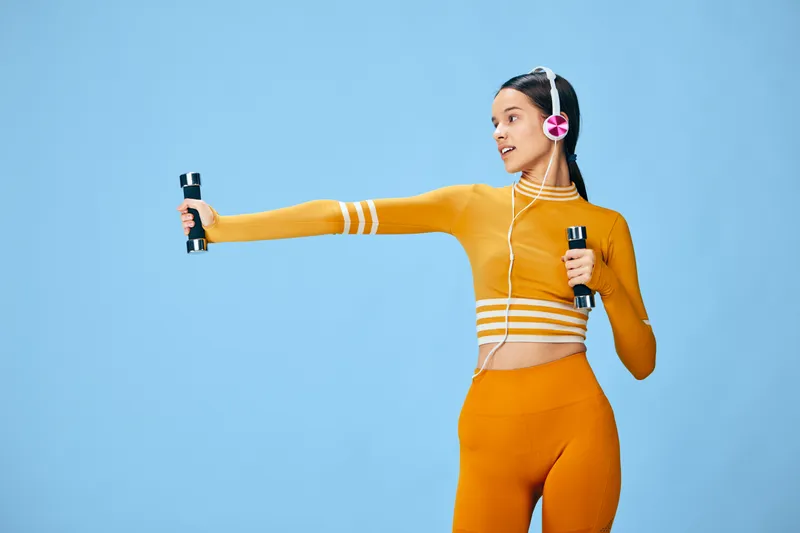 Female fitness athlete in orange sportswear holds dumbbells in each hand, performing a strength workout against a bright blue studio backdrop, wearing headphones, energetic, focused, and ready for