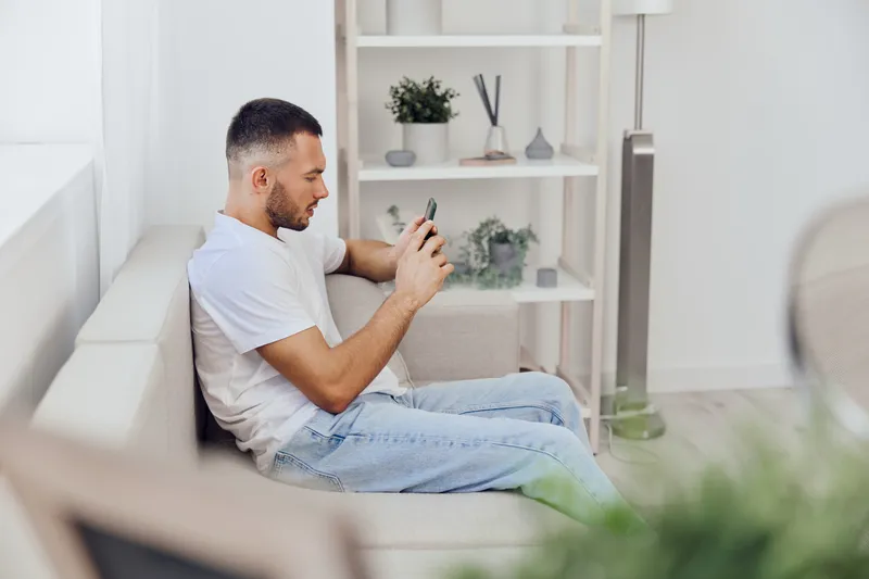 Thoughtful man using smartphone while relaxing at home, exploring digital detox and emotional minimalism in a soft and comfortable environment.