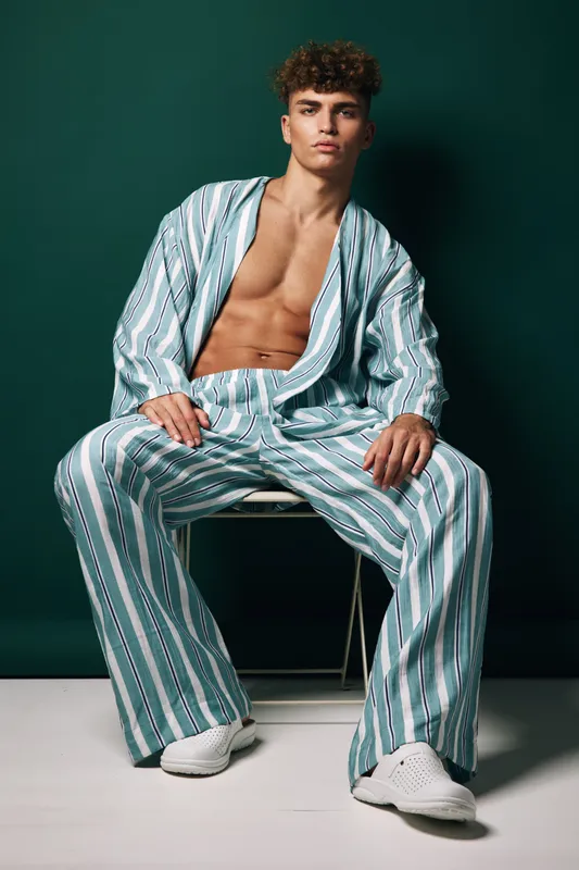 Young man with curly hair wearing striped loose pants and open shirt sitting on chair against dark background. Casual fashion, confident pose and relaxed style in studio.