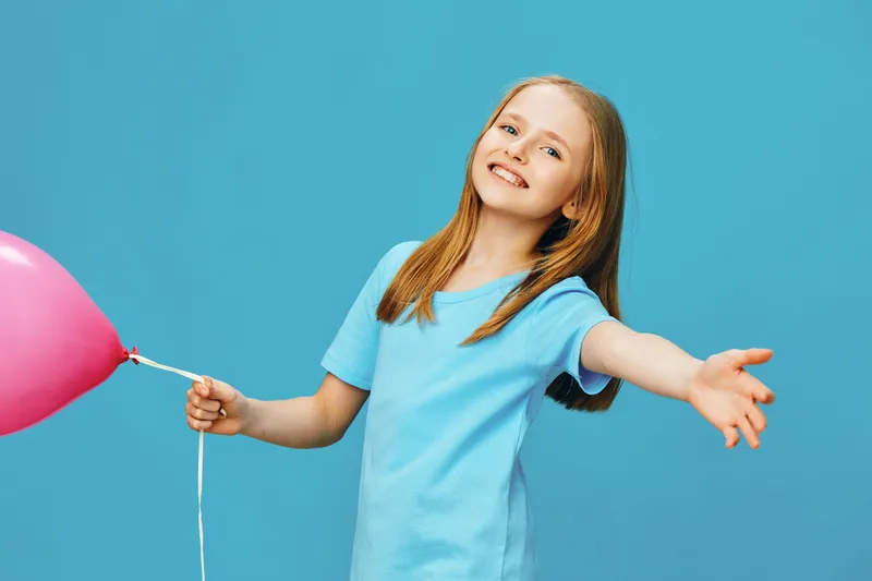 Happy girl with long red hair, wearing a light blue t shirt, joyfully holding a pink balloon against a bright blue background, embodying a cheerful childhood moment