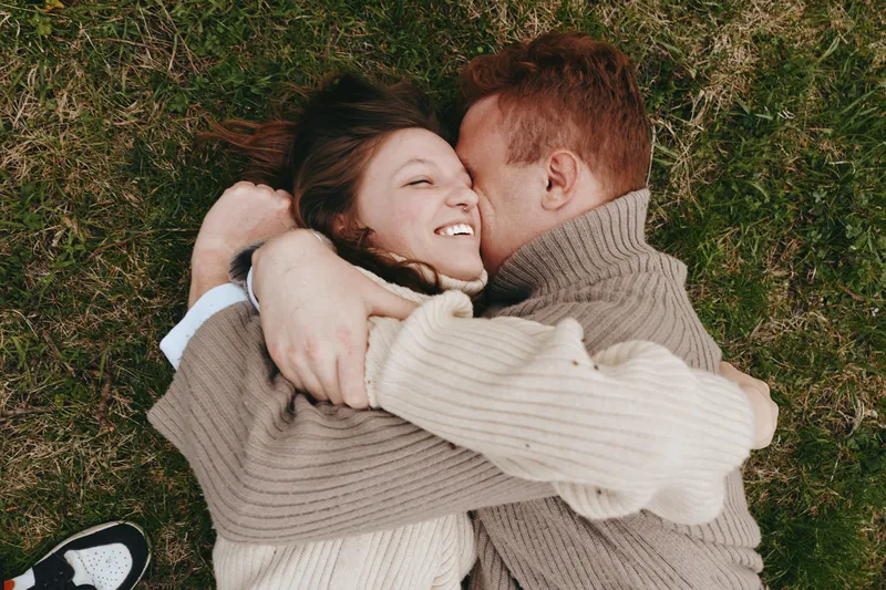 Romantic couple lying on grass, embracing and kissing, wearing knit sweaters, soft natural light, candid outdoor moment, warm cozy mood, lifestyle scene of affection and togetherness