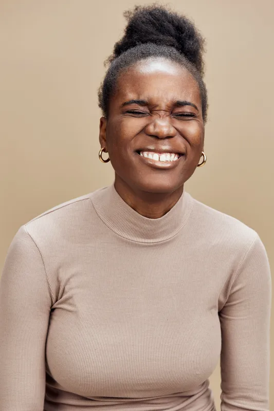 Cheerful young African woman smiling against a beige background, dressed in a light turtleneck, showcasing joy and happiness, ideal for conveying positivity and confidence