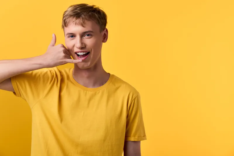 Smiling man in a yellow tee performs a call gesture against a yellow background, casual clothing and modern style for lifestyle branding, advertising and social media campaigns.