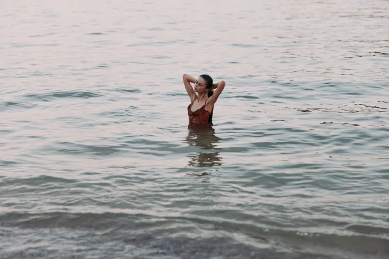 A lone swimmer stands waist deep in calm sea water with arms raised behind the head, wearing a dark swimsuit, capturing a serene, contemplative coastal moment at dusk.