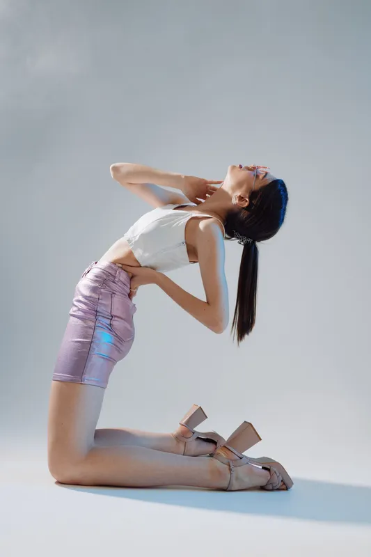 Young woman in a shiny pink skirt and white top poses on her knees with hands on hips and neck, showcasing beauty and fashion in a studio with soft lighting.