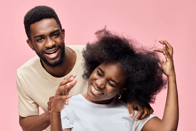 Happy diverse couple enjoying playful moment with vibrant hair in light clothing against pink background, showcasing joyful emotions and lively atmosphere