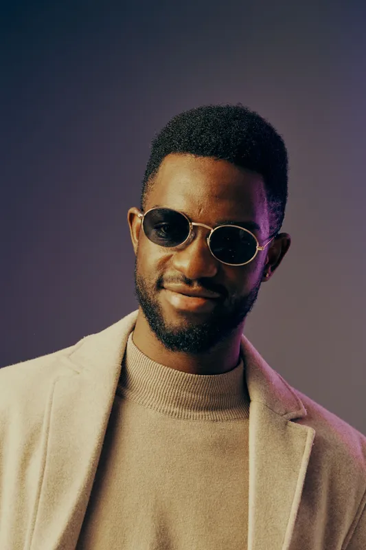Stylish young man in sunglasses, dressed in a beige coat and sweater, exuding confidence against a soft gradient background, showcasing modern fashion aesthetics