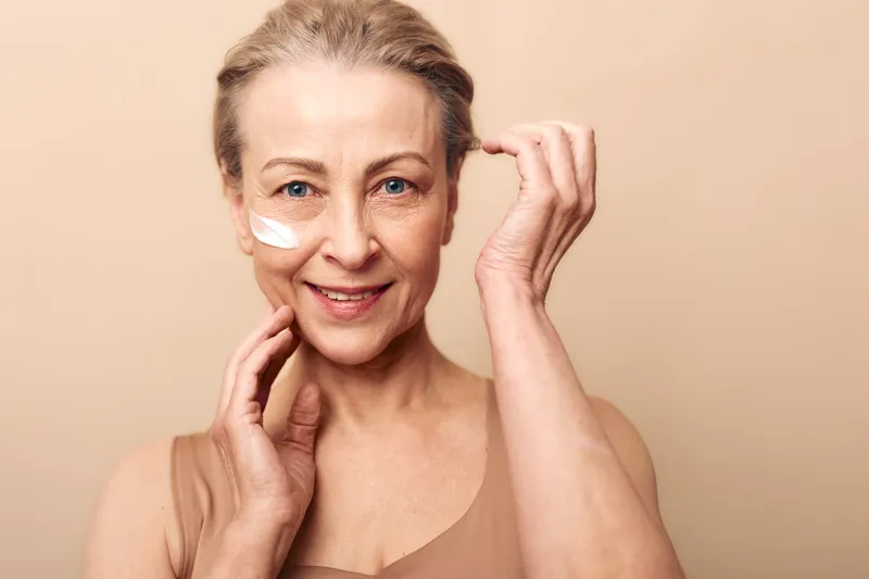 Smiling mature woman with natural skin applies white face cream on cheek. Close-up portrait on beige background. Concept of skincare, antiaging, selfcare, beauty, and wellness routine.