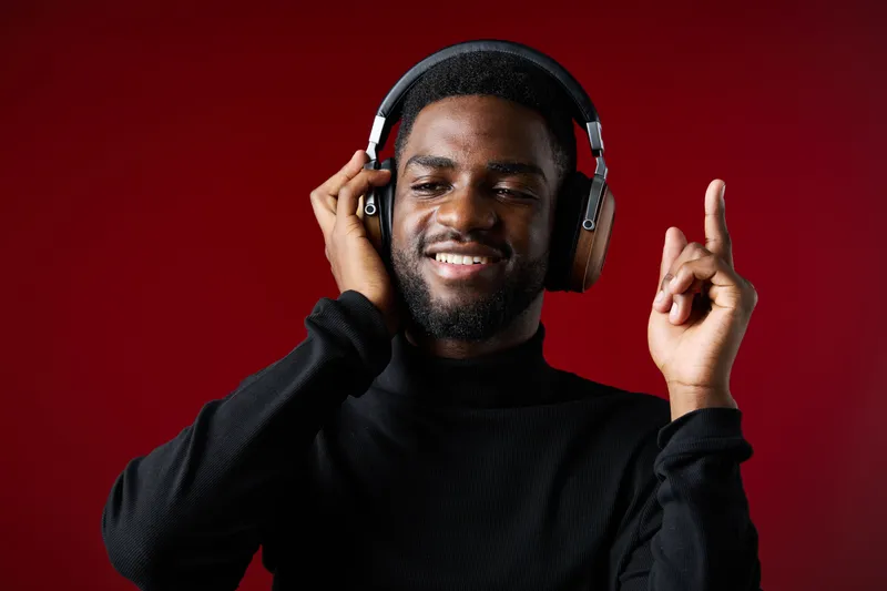 Young Black man enjoying music with headphones, smiling against a red background, showcasing modern style and vibrant emotions