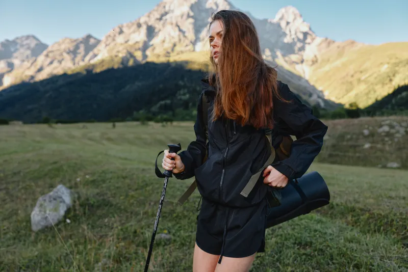 A confident hiker woman stands in a rugged mountain valley, gripping a trekking pole and wearing a waterproof jacket. She gazes ahead, ready for exploration in this bright alpine day.