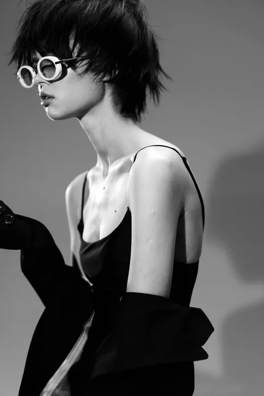 Black and white portrait of stylish woman wearing round glasses and fashionable dress with dark background.
