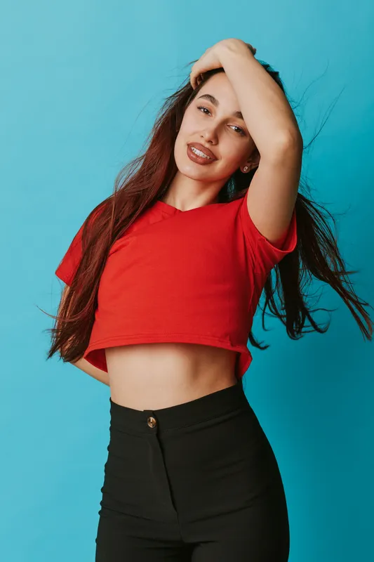Young woman posing with hand in hair wearing red cropped top and black pants against blue background. Fashion, beauty, casual style and confident female portrait studio shot.